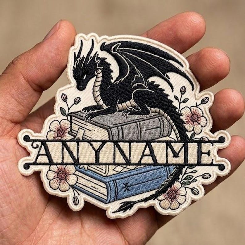 Op de afbeelding: Geborduurde patch met een zwarte draak op twee boeken en het woord "ANYNAME" eronder. De patch heeft een cr&egrave;mekleurige rand met roze en groene bloemendetails. De draak heeft gedetailleerde vleugels en schubben.