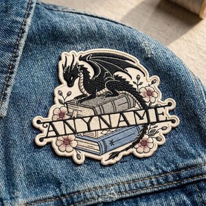 Op de afbeelding: Geborduurde patch met een zwarte draak op boeken, bloemendetails en de tekst "ANYNAME". De patch is geplaatst op een spijkerjasje. Het ontwerp combineert draken- en bloemenelementen.