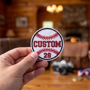 May include: A round baseball patch with a white background, red stitching, and the word "CUSTOM" in red block letters. The number "28" is below the word "CUSTOM". The patch is held in a hand.