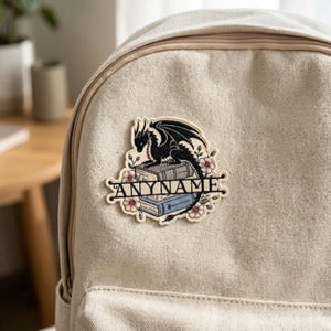 Op de afbeelding: Een zwarte drakenpatch met de tekst "ANYNAME" wordt getoond op een beige rugzak. De draak zit op twee boeken, omringd door roze bloemen. De patch is een decoratief accessoire.
