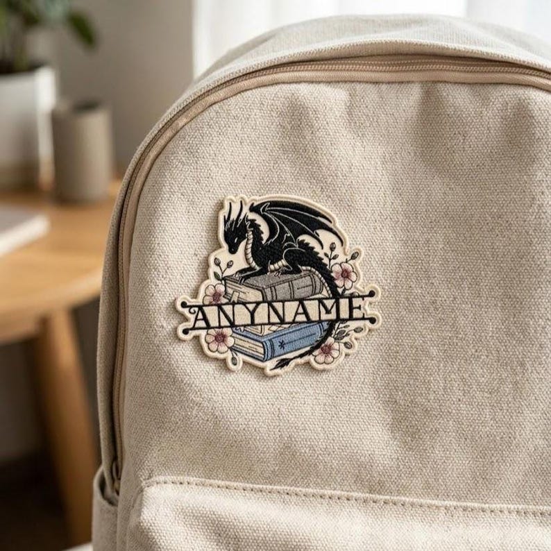 Op de afbeelding: Een zwarte drakenpatch met de tekst "ANYNAME" wordt getoond op een beige rugzak. De draak zit op twee boeken, omringd door roze bloemen. De patch is een decoratief accessoire.