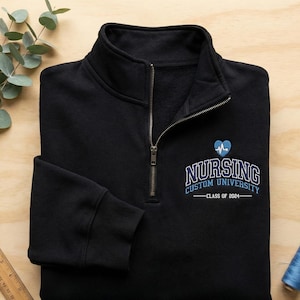 May include: Black quarter-zip sweatshirt with a stand-up collar. Features a zipper closure and ribbed cuffs. The sweatshirt has the text "NURSING CUSTOM UNIVERSITY" with a heart and the text "CLASS OF 2024" in blue.