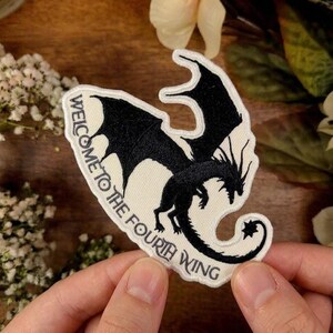 May include: A black dragon patch with the text "WELCOME TO THE FOURTH WING" embroidered on a cream-colored background. The dragon has detailed wings and a curled tail. The patch is held in a person's hands.