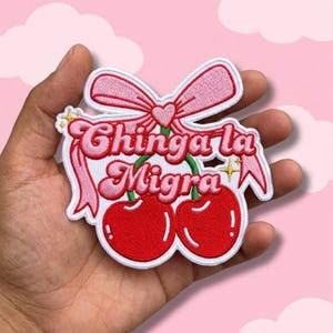 May include: Embroidered patch featuring two red cherries with green stems, a pink bow, and the text "Chinga la Migra" in pink script. The patch has a white border and is set against a pink background.