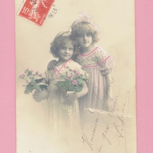 Può includere: Cartolina vintage con due bambine in abito, una con fiori. Un francobollo francese è nell'angolo in alto a sinistra. La cartolina presenta scritte a mano e una palette di colori tenui e sbiaditi.