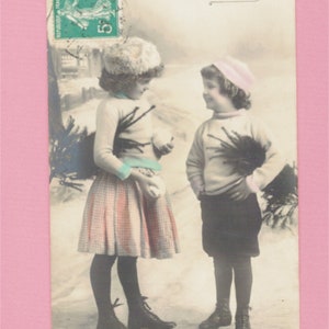 May include: A vintage postcard featuring two children in winter clothing. The girl on the left is holding a snowball and the boy on the right has his hands in his pockets. The postcard has the text "Joyeux Noel" written on it.