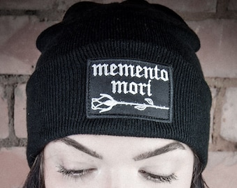 Pode incluir: Um gorro preto com uma rosa bordada branca e o texto "memento mori" na frente.
