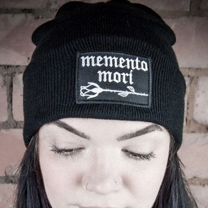 May include: A black beanie with a white embroidered rose and the text "memento mori" on the front.