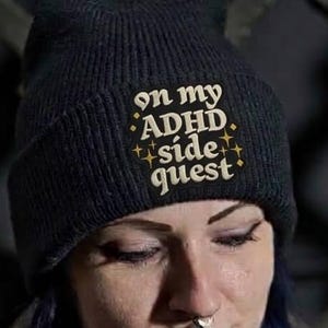 May include: A black knit beanie featuring the embroidered text "on my ADHD side quest" in a light tan color, accented by small star symbols. The beanie is made of a ribbed fabric and is worn on the head, showcasing the clear, centered lettering.