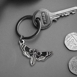 May include: A silver key chain with a black and white moth charm, a silver key with the word "TWIC" on it, and a silver ring. The keychain is on a gray surface with two silver coins.