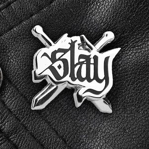 May include: A silver-toned metal pin featuring the word Slay in a stylised black font, layered over two crossed swords. The pin is attached to a black leather garment with a visible circular metal snap button near the left edge of the frame.