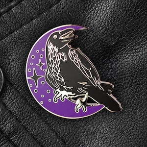 Puede incluir: Pin de esmalte morado con un cuervo negro posado en una luna creciente. La luna tiene un diseño estrellado.