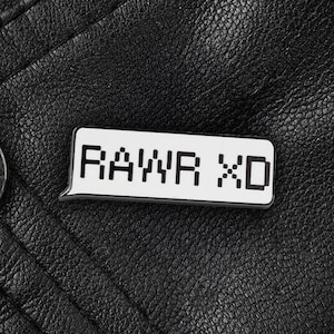 Op de afbeelding: Een witte emaille pin met zwarte tekst die "RAYVR XD" luidt op een zwarte lederen achtergrond.