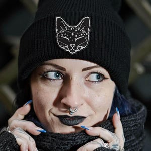 May include: Black beanie hat with a white cat face design and stars. The person in the image is wearing a black scarf and gloves. They have blue eyes, black lipstick, and blue nail polish.