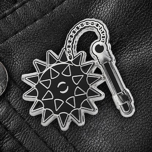May include: A silver-colored metal pin shaped like a spiked ball on a chain. The ball has a black center with a white circle and a white letter C in the center.