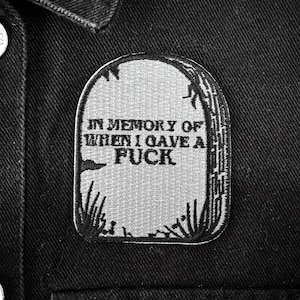 May include: A gray patch in the shape of a tombstone with the words "IN MEMORY OF WHEN I GAVE A FUCK" in black lettering. The patch is sewn onto a black denim jacket. The tombstone design includes grass and a decorative border.