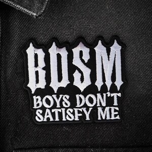 May include: Black embroidered patch with the text "BDSM BOYS DON'T SATISFY ME" in white gothic lettering. The patch is sewn onto a black denim jacket, with a button visible.