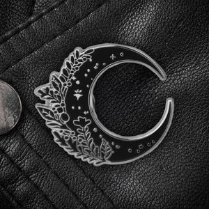 Puede incluir: Un broche plateado con forma de media luna con relleno de esmalte negro. El broche presenta detalles florales y de estrellas. El broche está sobre un fondo de cuero negro, con una moneda visible.
