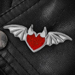 Puede incluir: Un emblema plateado en forma de corazón con alas de murciélago y un centro rojo. El emblema está colocado sobre una superficie de cuero negro. A la izquierda del emblema se ve una moneda.