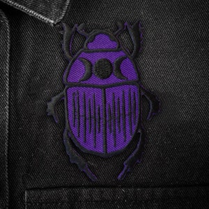 Puede incluir: Parche bordado morado con forma de escarabajo, con detalles negros y fases lunares. Cosido sobre una chaqueta vaquera negra, con botones plateados. Un accesorio único para personalizar ropa y bolsos.