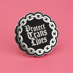 May include: Black and silver enamel pin with a chain border and the text "Protect Trans Lives" in a gothic font.