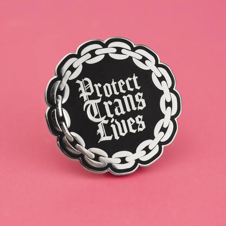 May include: Black and silver enamel pin with a chain border and the text "Protect Trans Lives" in a gothic font.