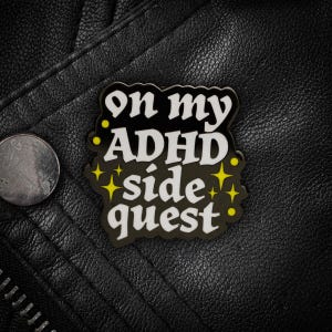 May include: A black enamel pin with a scalloped edge featuring the white text "on my ADHD side quest" and small yellow star accents. The pin has a glossy finish and is shaped to follow the contour of the bold, stylized lettering.