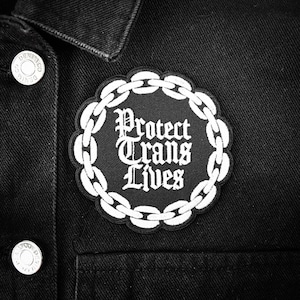 Op de afbeelding: Zwart-witte patch met een kettingrand en de tekst "Protect Trans Lives" in een gotisch lettertype.