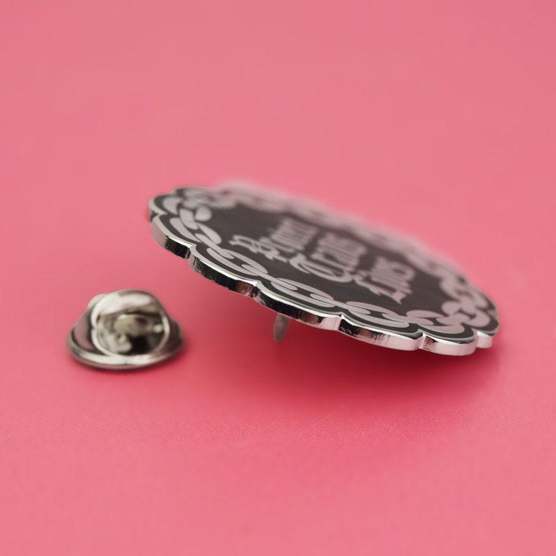 May include: Silver pin with a black background and white text that reads "Funny Cat Lady". The pin has a decorative border.