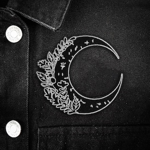 May include: A white embroidered crescent moon patch, adorned with floral and star motifs, attached to a black denim jacket. The patch's design includes leaves, flowers, and stars, offering a celestial and botanical aesthetic.