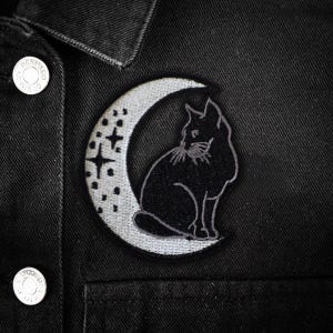 Könnte beinhalten: Schwarzer Katzen-Patch auf einem schwarzen Jeansjacken-Stoff. Der Patch zeigt eine schwarze Katze, die in einem Halbmond mit Sternen sitzt. Der Mond ist hellgrau mit kleinen Punkten.