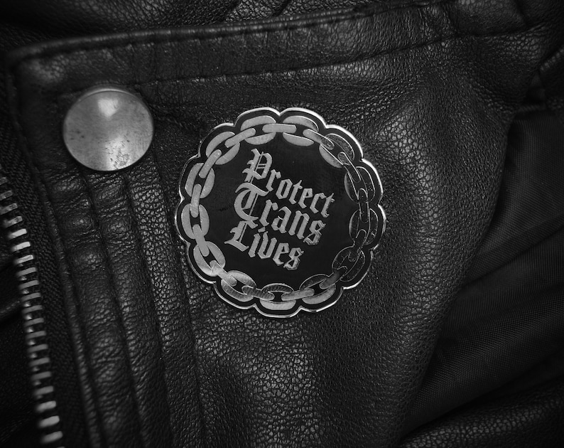 May include: A black leather jacket with a silver pin that says "Protect Trans Lives" in a gothic font.