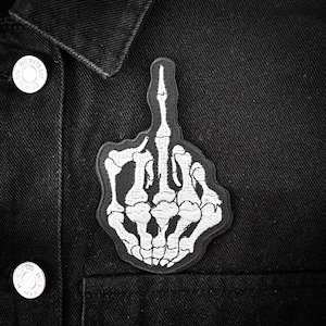 Könnte beinhalten: Schwarze Jeansjacke mit einem schwarz-weißen, gestickten Aufnäher einer Skeletthand, die den Mittelfinger zeigt. Der Aufnäher befindet sich auf der Vorderseite der Jacke, in der Nähe der Tasche.
