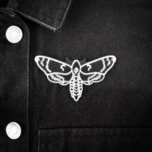 Könnte beinhalten: Eine schwarze Jeansjacke mit einem weißen, gestickten Motten-Patch auf der Vorderseite. Die Motte hat ein Totenkopf- und Knochenkreuz-Design.