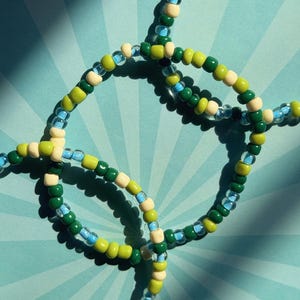 May include: Close-up of beaded jewelry featuring a pattern of green, light green, blue, and off-white beads. The beads are strung together to form a circular design. The background is a light blue radial pattern.