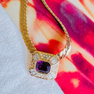 May include: A gold chain necklace with a pendant featuring a large, square-shaped purple gemstone surrounded by smaller clear gemstones.