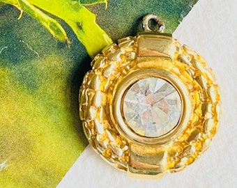May include: A gold pendant with a clear rhinestone in the center. The pendant is surrounded by a decorative gold chain.