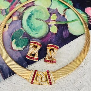 May include: A gold-toned omega chain necklace and matching clip-on earrings set. The jewelry features a central interlocking link design accented with rectangular red stones and small clear pavé-set crystals along the edges of the metal links.