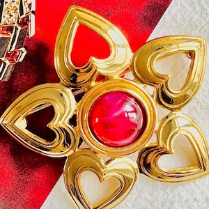 May include: A gold-toned brooch with a flower design, featuring heart-shaped petals surrounding a central red gemstone. A gold necklace with black and clear accents is partially visible in the background. The brooch is on a red and white background.