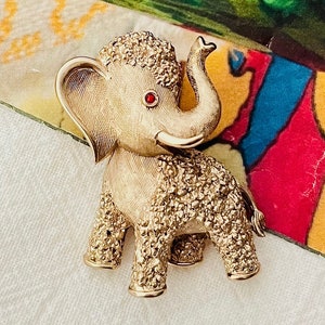 May include: A gold-tone elephant brooch with red rhinestone eyes. The elephant is standing on all four legs with its trunk raised. The brooch is textured and has a sparkly finish.