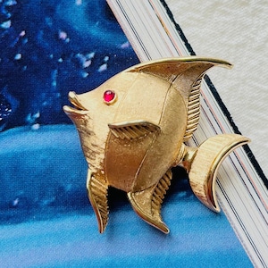 Op de afbeelding: Een gouden vis broche met een rode strass steentje oog. De broche ligt op een blauw en wit boek.