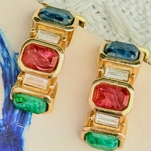 May include: A pair of gold-tone hoop earrings featuring a multi-colored design. Each earring is set with rectangular-cut stones in alternating colors of deep blue, clear white, vibrant red, and emerald green, arranged in a vertical row.