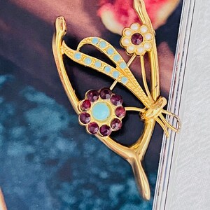 Könnte beinhalten: Eine goldene Brosche in Form eines Schmetterlings mit blauen und weißen Emaille-Akzenten und roten und türkisfarbenen Edelsteinen.