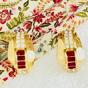 May include: A pair of gold hoop earrings with red and clear rhinestones. The earrings are on a floral patterned fabric.