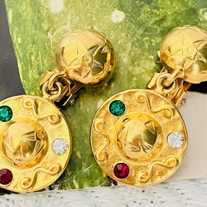 Puede incluir: Un par de pendientes dorados de clip. Cada pendiente presenta un colgante redondo y decorativo con un diseño central y tres piedras preciosas de colores: verde, rojo y blanco. Los pendientes tienen un diseño texturizado de estrella en la parte superior.