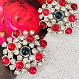 May include: Two vintage silver-toned clip-on earrings. Each earring features a floral design with red and clear rhinestones, and a single dark blue center stone. The earrings are set against a backdrop of red roses.