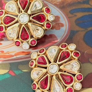 May include: A pair of gold-tone clip-on earrings featuring a central clear round stone surrounded by alternating teardrop-shaped red and clear stones, with additional small red and clear round stones set along the outer edges of the design.