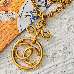 May include: A gold-coloured necklace with a large, round pendant featuring interlocking C's within a circle. The necklace has a thick chain and a bow-shaped connector. The jewellery is displayed on a patterned surface.