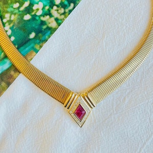 May include: A gold chain necklace with a red gemstone pendant surrounded by clear rhinestones. The necklace is on a white surface.