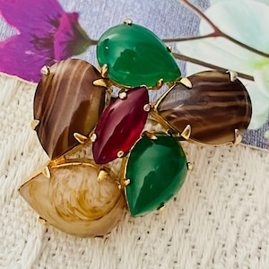 May include: A cluster of colourful, faceted stones set in gold metal. The stones are green, brown, and red. The brooch is a unique and eye-catching piece of jewellery.